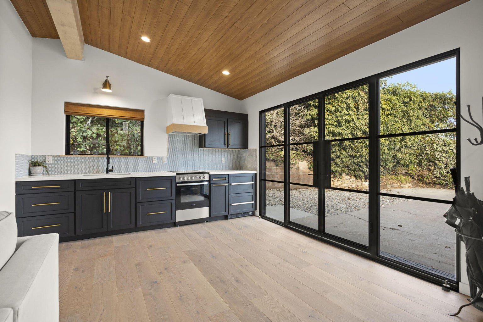 A modern kitchen with dark cabinets, stainless steel appliances, light wood flooring, and a wood ceiling. Sliding glass doors open to a patio area with greenery outside.